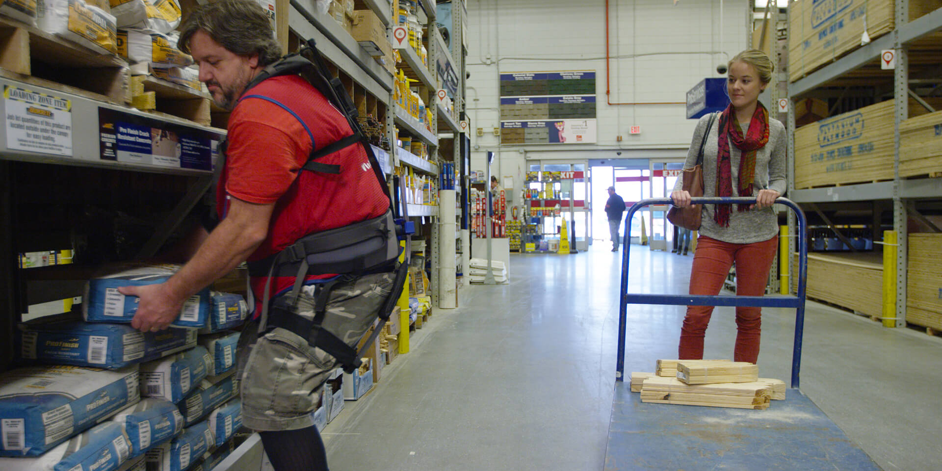 Lowe's Home Improvement associate on sales floor lifting heavy object for female customer while wearing soft robotic exoskeleton for assistance.